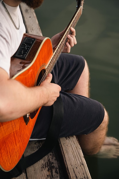 Een man speelt gitaar zittend op een houten pier in de buurt van het meer, het concept van de zomer buiten