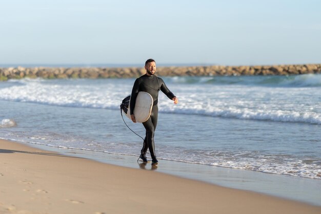Een man surfer in een wetsuit die op de kust loopt en een surfplank vasthoudt