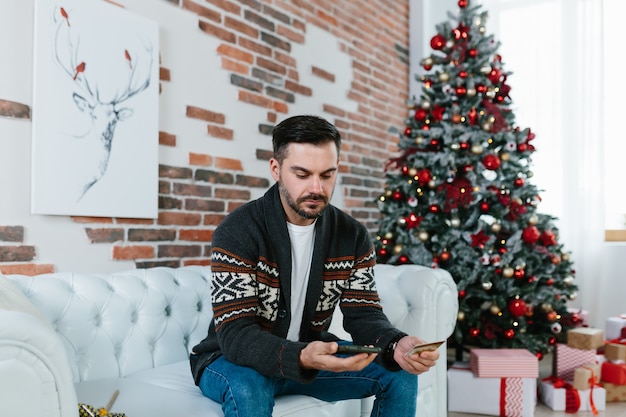 Een man thuis doet aankopen, gebruikt een creditcard en een smartphone, kiest cadeaus voor familie en vrienden voor Kerstmis