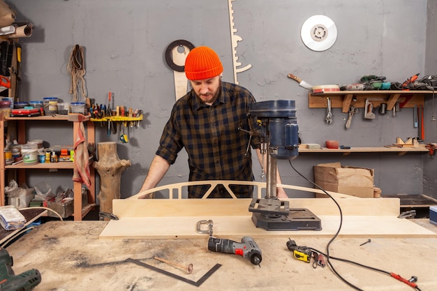 Een man timmerman in een hoed en een shirt snijdt een houten plank op een grote boormachine in een zijaanzicht van een werkplaats op de achtergrond veel gereedschap