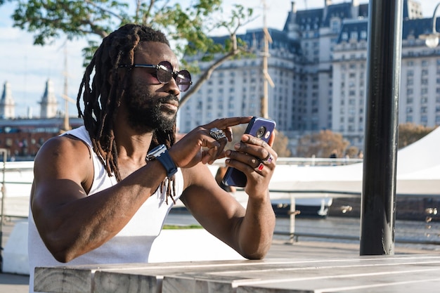 Een man van Afrikaanse afkomst met dreadlocks die buiten zit en het scherm van zijn mobiele telefoon aanraakt en op het internet surft.