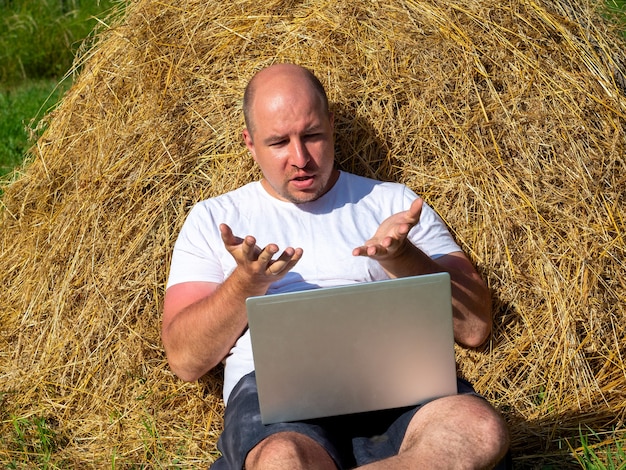 Een man van middelbare leeftijd, gekleed in een T-shirt en korte broek, zit bij een hooiberg met een laptop en houdt een vergadering. Werken op afstand, emoties, platteland