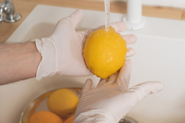 Een man wast een citroen met een wasmiddel in handschoenen. Een jonge man steriliseert groenten en fruit tijdens een epidemie. Handen in wegwerphandschoenen close-up was een citroen onder stromend water.