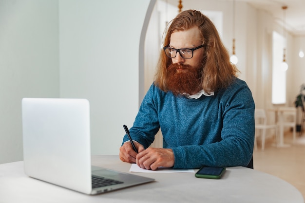 Een man werkt op een laptop in een café en schrijft een notitie op papier