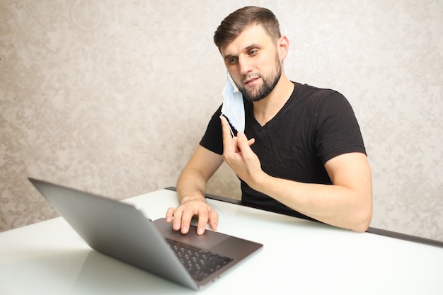 een man werkt thuis in isolatie op een laptop en heeft een medisch masker over zijn oor hangen