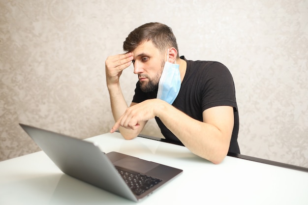 een man werkt thuis in isolatie op een laptop en heeft een medisch masker over zijn oor hangen