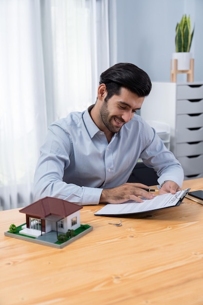 Een man zit aan een bureau met een huisje en een klein model van een huisje.