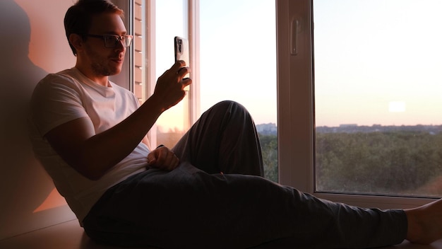 Een man zit bij de raamtelefoon en kletst op de zonsondergang glimlach en maakt foto's van zonsondergang