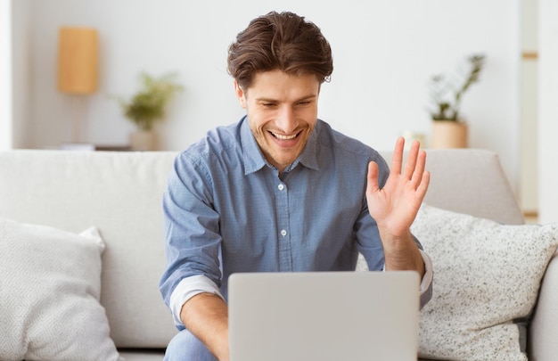 Een man zit op een bank glimlachend en zwaait naar het laptop scherm voor hem