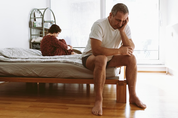 Een man zit op een bed met een vrouw op de achtergrond.