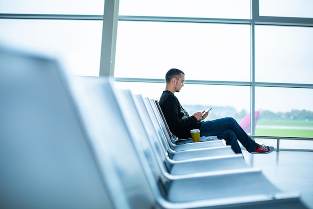 Een man zit op een lege rij stoelen voor een groot glas-in-loodraam in een luchthaventerminal, wachtend op een vlucht