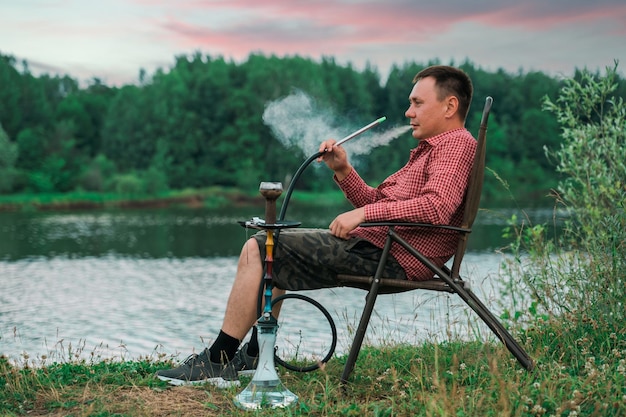 Een man zit op een stoel aan de rivieroever en rookt een waterpijp