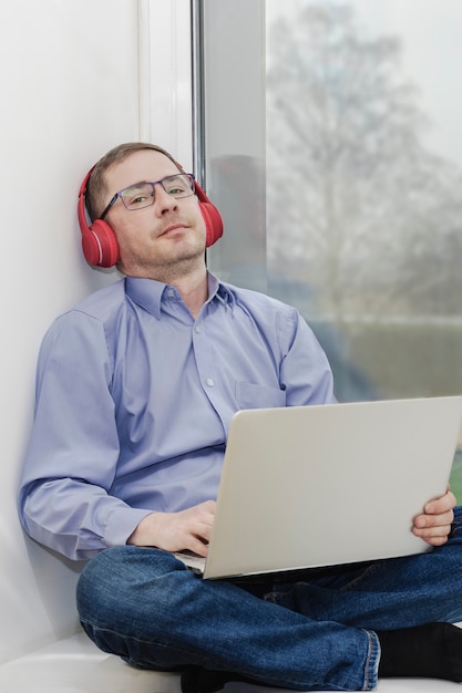 Een man zit op een vensterbank in een koptelefoon met een laptop ontspanning en luisteren naar muziek