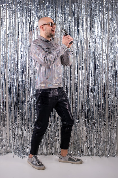 Een man zonder haar met zonnebril in een glanzend overhemd, een zwarte leren broek en grijze sneakers zingt in een kleine zilveren microfoon.