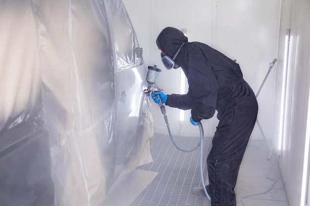 Een mannelijke arbeider in jumpsuit en blauwe handschoenen schildert met een spuitpistool een zijdeel van de carrosserie in het zwart na beschadigd te zijn geraakt bij een ongeval Beroepen in de autoservice-industrie