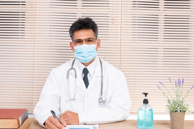 Een mannelijke arts, die een masker en witte zelfklevende kleren draagt, zit aan zijn bureau.