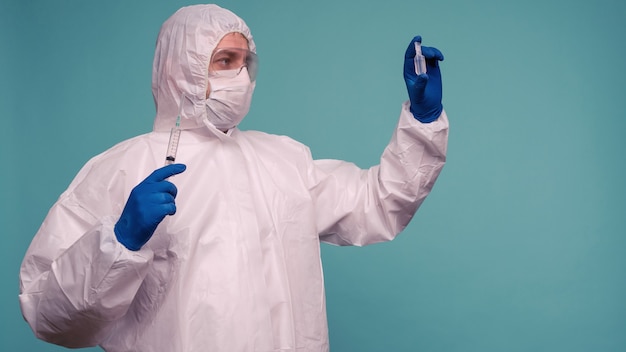 Een mannelijke arts in beschermende overall en een masker houdt een ampul met een vaccin en een spuit in zijn handen.