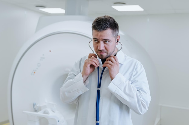 Een mannelijke arts in een witte jas met een stethoscoop in zijn handen tegen de achtergrond van een moderne ct-scanner in de kliniek