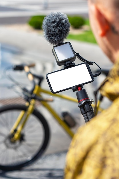 Een mannelijke blogger houdt een statief in zijn handen met een smartphonemodel met een wit scherm en een lampje met een microfoon op de achtergrond van een fiets