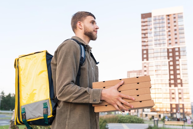 Een mannelijke koerier met een gele thermoszak houdt pizzadozen vast tijdens de levering aan een klant tegen de achtergrond van gebouwen