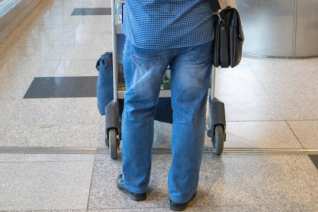 Een mannelijke passagier in een spijkerbroek en een shirt die een karretje met bagage en handbagage duwt op het luchthavenclose-up achteraanzicht reisconcept