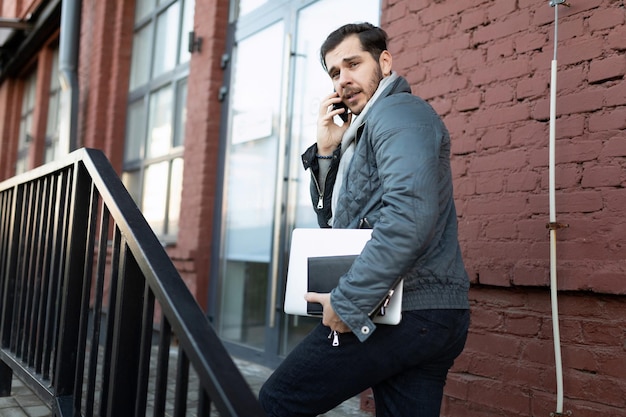 Een mannelijke programmeur pratend aan de telefoon met een laptop in zijn handen komt het kantoorgebouw binnen
