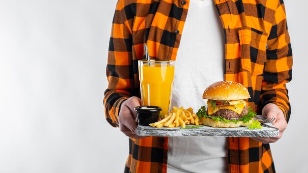 Foto een mannelijke student in een geruite oranje hemd op een witte achtergrond houdt een houten bord vast met een verse burger een glas sinaasappelsap en gebakken aardappelen met kopieerruimte voor tekst