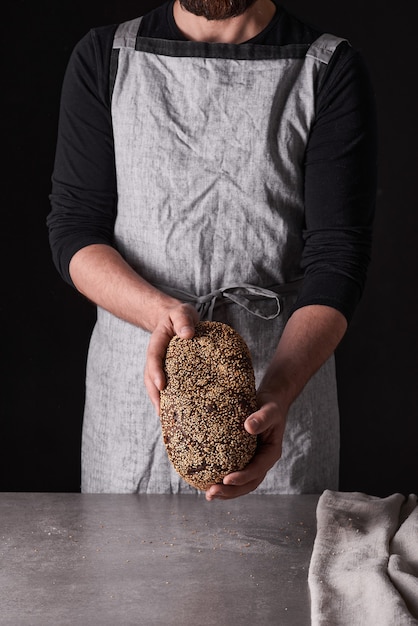 Een mannenbakker met een baard in een grijs schort staat tegen een zwarte achtergrond en houdt, breekt, snijdt heerlijk, knapperig brood, broodjes, stokbrood.