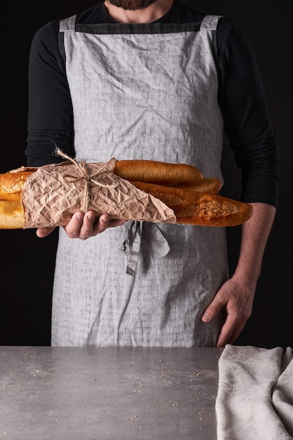 Een mannenbakker met een baard in een grijs schort staat tegen een zwarte achtergrond en houdt, breekt, snijdt heerlijk, knapperig brood, broodjes, stokbrood.