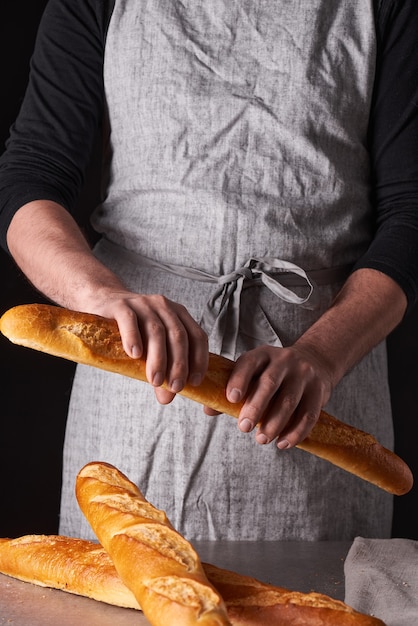 Een mannenbakker met een baard in een grijs schort staat tegen een zwarte achtergrond en houdt, breekt, snijdt heerlijk, knapperig brood, broodjes, stokbrood.