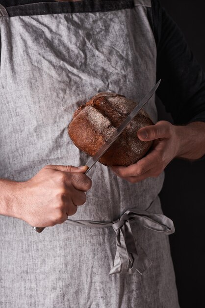 Een mannenbakker met een baard in een grijs schort staat tegen een zwarte achtergrond en houdt, breekt, snijdt heerlijk, knapperig brood, broodjes, stokbrood.