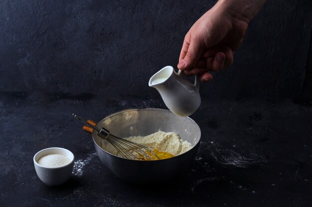 Een mannenhand giet melk van een melkboer in een metalen kom om deeg te maken. Ingrediënten en gebruiksvoorwerpen voor het koken van cake op donkere tafel. Het concept van het maken van deeg voor het bakken