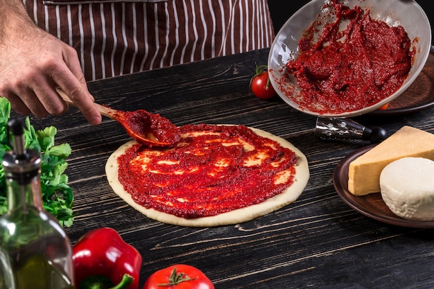 Een mannenhand verspreiding van tomatenpuree op een pizzabodem met lepel op een oude houten achtergrond. Koken concept. Detailopname