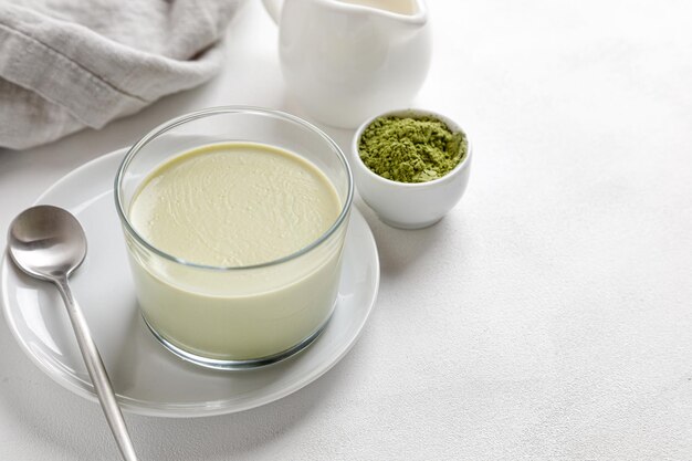 Een matcha thee panna cotta dessert in een glas met lepel op een witte achtergrond en een grijs servet