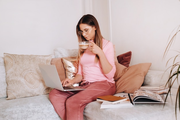 Een meisje dat 's ochtends in pyjama's thuis werkt aan een laptop met koffie drinken, een meisje dat zichzelf thuis isoleert en op de bank rust en naar een laptop kijkt. Huishoudelijke klusjes.