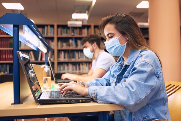 Een meisje en een jongen met maskers studeren met laptops in de bibliotheek