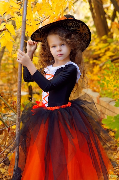 een meisje gekleed als een kleine heks in een oranje rok en een puntige zwarte hoed met een bezem in een herfst Halloween Park