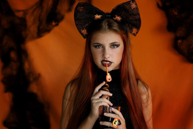Een meisje gekleed voor Halloween houdt een glas met een rietje in haar handen op een oranje achtergrond