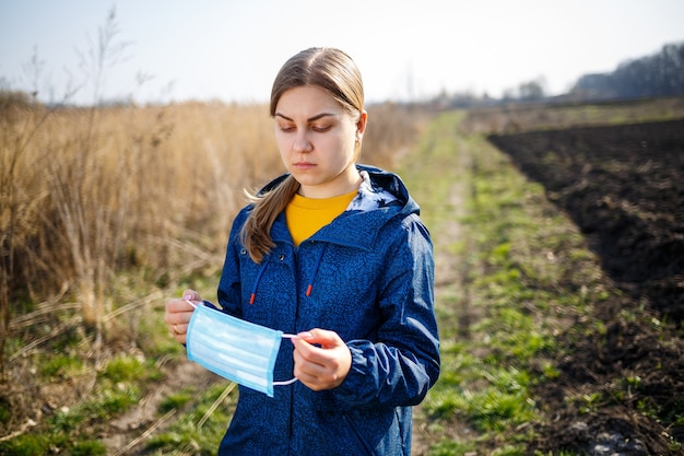 Een meisje in een blauwe jas op de natuur houdt een beschermend masker in haar handen. Zonnige dag buitenshuis masker op het gezicht van het virus. virale ziekte