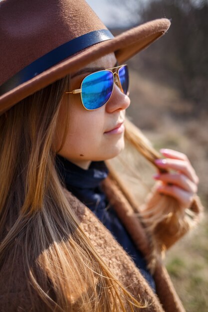 Foto een meisje in een bruine jas, hoed en bril loopt in het park met een meer onder de felle zon.