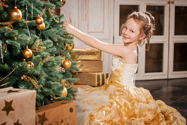 Een meisje in een elegante jurk hangt decoraties aan de kerstboom.