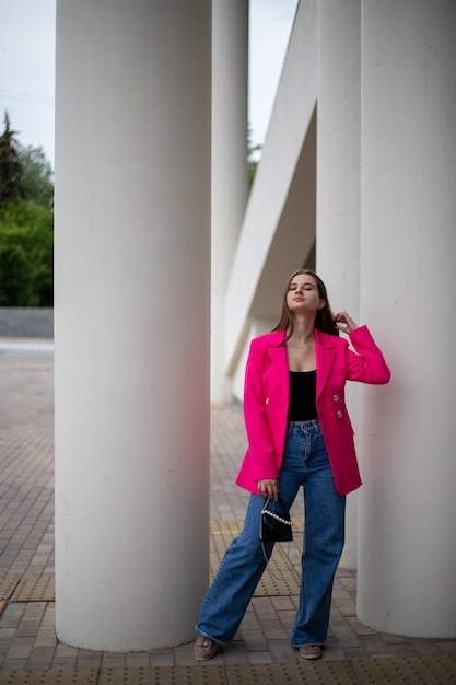 een meisje in een fel fuchsiakleurig jasje en een spijkerbroek staat lang op straat met een kleine handtas