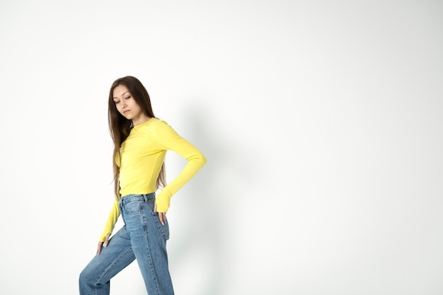 Een meisje in een geel topje en spijkerbroek staat voor een witte muur.