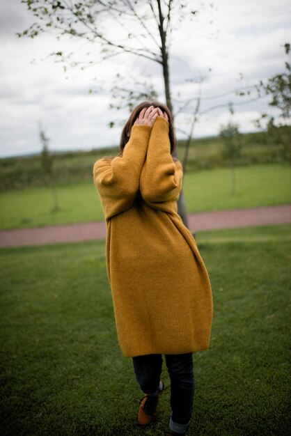 Een meisje in een gele jas bedekt haar gezicht met haar handen, staande in het park