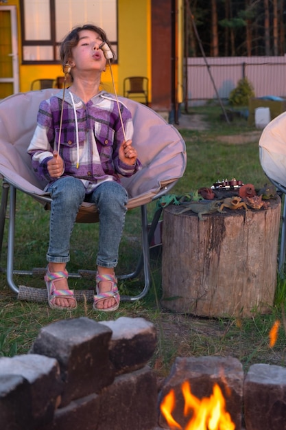 Een meisje in een geruite shirt roostert marshmallows op een vuur in de tuin van het huis avond familie verzamelt zich bij het kampvuur buiten toeristische stoelen