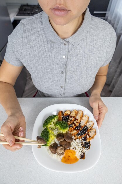 Een meisje in een grijs shirt in de keuken thuis houdt een bord gouden rijst shiitake champignons kip met unagi saus en broccoli in haar handen Aziatisch eten concept