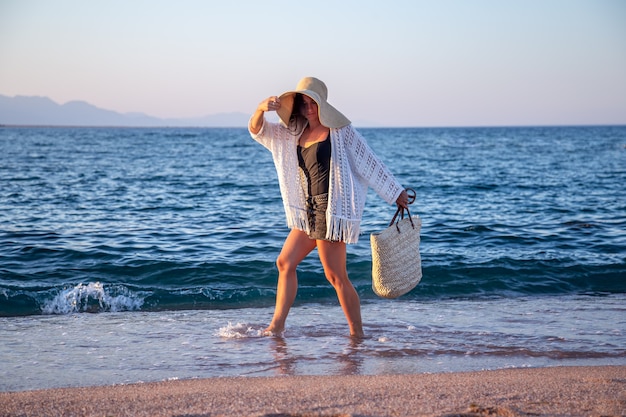 Een meisje in een grote hoed met een rieten tas loopt aan de kust. Zomer vakantie concept.