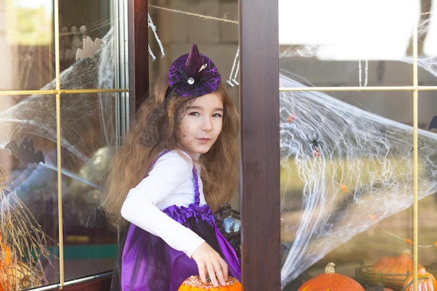 Een meisje in een heksenkostuum op de vensterbank bij het raam speelt met een spinnenweb en een spin die het huis decoreert voor de halloween-vakantie vreselijke landschappen, angst en horrorpompoenen