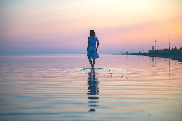 Een meisje in een jurk aan de kust bij zonsondergang loopt over het water