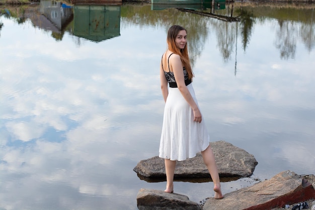 Een meisje in een jurk op een steen in de rivier met een weerspiegeling van de lucht met wolken in het water